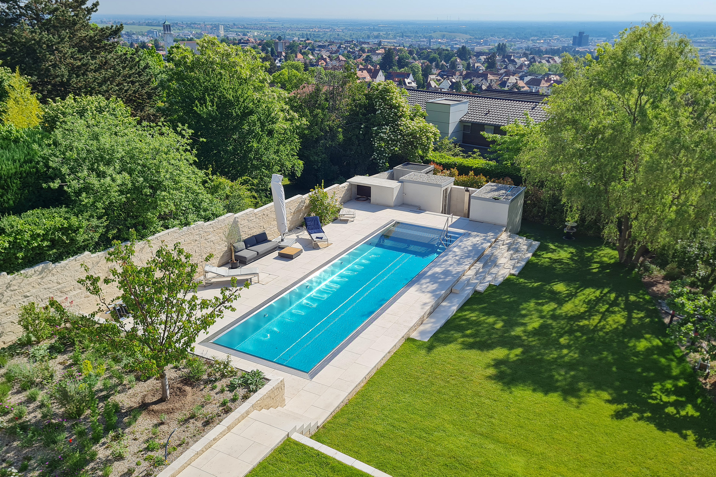 Edelstahlschwimmbecken mit Unterflurabdeckung und weitläufigem Blick über ein Wohngebiet, eingebettet in Hanglage